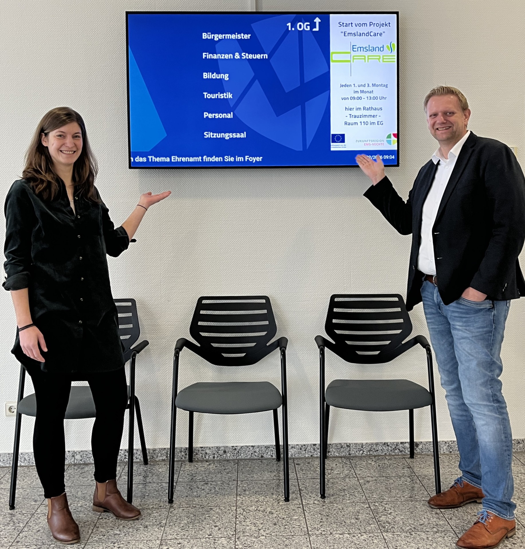 Bild von der Beraterin Frau Schulten mit dem Samtgemeindebürgermeister Matthias Lühn vor dem Fernseher im Foyer, auf dem das Projekt "EmslandCare" beworben wird