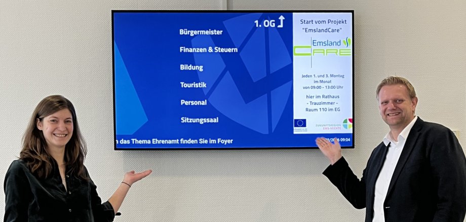 Bild von der Beraterin Frau Schulten mit dem Samtgemeindebürgermeister Matthias Lühn vor dem Fernseher im Foyer, auf dem das Projekt "EmslandCare" beworben wird